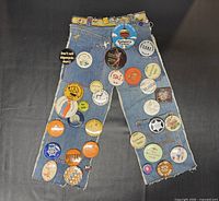 Denim pant leg shaped pin/button display with assorted vintage pins/buttons attached showing color and condition.