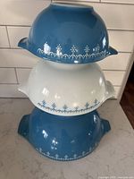 Stacked view of three Cinderella nesting bowls showing snowflake garland pattern around the exterior. Two blue, one white bowl. Side handles visible.
