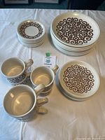 Photo showing multiple stacked dinner plates, lunch plates, side plates, and four cups with brown decorative pattern on cream ceramic base.