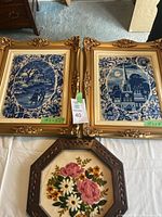Two matching blue and white crewel needlepoints in ornate gold frames, and one floral crewel needlepoint in a wooden octagonal frame, all displayed on a white sheet.