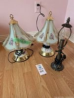 Set of 3 lamps on wooden floor, two have floral glass shades with brass base, one smaller with dark metal base and missing shade