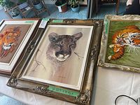 Three animal artworks displayed on a table: a Phil Prentice cougar painting in a large ornate frame, a lion crewel embroidery in a plain frame, and a tigers crewel embroidery in a gold frame.