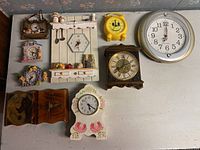 Nine assorted clocks arranged on a white table against a wall with floral wallpaper, showing a variety of decorative themes and materials such as wood, plastic, and ceramic. Clocks include hexagonal, round, and house shapes with visible scratches and wear.
