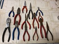 Ten assorted older hand pliers arranged on tiled floor, varied handle colors and sizes.