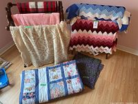 View of all blankets and tablecloths including folded patchwork quilt, crocheted beige blanket, red and black checkered blanket on wooden blanket stand, multicolored zigzag crochet blanket, pink plush blanket, and two dark textured blankets.