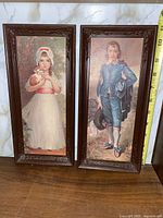 Pair of vintage paintings showing a girl holding a small animal and a boy in blue historical costume, framed in carved wooden frames.