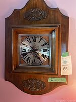 Full view of wooden framed Caravelle wall clock with decorative carvings above and below the clock face; rectangular silver-tone dial with black Roman numerals and ornate black hands.