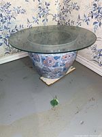 Side view of ceramic planter with floral pattern and round glass top on basement floor with floral wallpaper background.