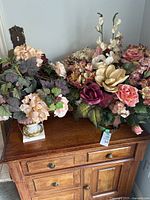 Two artificial floral arrangements on a wooden cabinet, larger arrangement to the right and smaller arrangement to the left