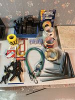 Various hanging supplies on table including black plastic plate holders, clamps, adhesive tapes, bungee cords, metal brackets, and small hardware items.
