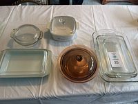 Full view of all 12 cookware pieces arranged on a table: 4 clear glass baking dishes (various shapes), 1 amber glass round casserole with lid, 1 white ceramic casserole dish with floral pattern and lid.