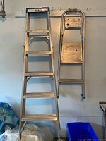 Two ladders hanging on wall: tall 6 ft aluminum step ladder with wide steps and smaller folding ladder with two steps and flat platform.