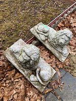 Pair of weathered concrete lion statues on leafy ground next to moss, positioned outdoors.
