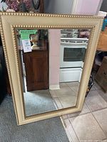Full view of the mirror showing the rectangular shape with a gold beaded frame, placed leaning against a wall on carpet and tile.