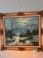 Full view of the framed landscape painting showing winter scene with cabin, snow, mountains, and trees.