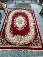 Full view of vintage red area rug with cream floral oval border and central medallion, showing fringe and some wear spots.