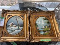 Both framed oval landscape paintings displayed on a glass table, showing ornate gold wooden frames with detailed corners and floral accents