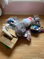 Wide view showing multiple bags and boxes of craft supplies on wooden floor, including yarn skeins, plastic canvas, and a box containing thread and needles