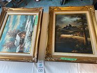 Two framed landscape paintings placed on a table, one to the left with a snowy winter cabin scene signed Jankowski, one on the right with a dark mountain and cabin scene signed R Carson.