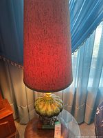 Full view of the lamp showing the tall cylindrical reddish-brown speckled fabric lampshade over an ornate green and gold base with silver pedestal, placed on a round wooden table.