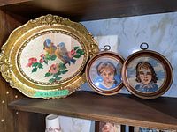 Three vintage needlepoints on shelf with one oval ornate gold frame and two round wooden frames