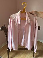 Light pink lightweight jacket hanging on hanger held by hand, shown from front