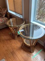 Two wicker and rattan side tables with round clear glass tops, positioned beside a window on wood flooring, showing overall condition and design.