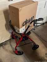 Photo of Drive brand fold-up walker in red with black handles and wheels, with boxed item placed behind it.