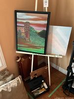 Image of wooden standing easel with framed painting and blank canvas behind it. Black plastic case with brushes and painting knife at base of easel.
