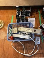 Power strip with multiple sockets, cords, and adapters in basket with phones and base unit in background