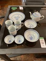 Full set displayed on glass table showing all 9 pieces including teapot, cake plate, mugs, medium and small bowls.