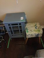 Blue painted cabinet with wood framed glass door and adjustable inside shelf, standing next to floral patterned upholstered footstool with wooden legs.
