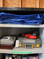Shelf containing gray plastic carrying case for Mastercraft cordless drill with accessories, and a black and red corded power tool in a plastic storage case. Large blue folded tarp placed on the top shelf.