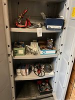 Tools and hardware arranged on four shelves inside a grey cabinet including clamps, wires, metal tools, and boxes containing small parts.
