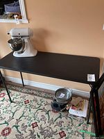 Black rectangular fold-up table with a white kitchen stand mixer and other items beneath. Shows minor wear on top surface.