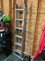 Full view of the folded metal ladder resting vertically against a wood paneled wall in a storage area.