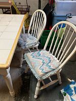 Four white Windsor style kitchen chairs with spindle backs, turned legs, minor scratches, and reversible cushions with zigzag fabric pattern