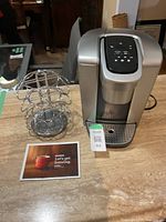 Keurig coffee maker next to metal coffee pod holder and a Keurig brochure on table.