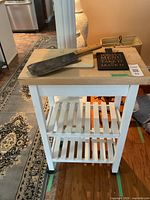 Front view of wood top kitchen cart painted white, showing two lower slatted shelves on casters with cleaver, cutting board and sign on top.