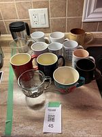Wide view of 13 kitchen mugs arranged on a countertop including a black travel mug and ceramic mugs in white, red, blue, floral, vintage print, and brown colors.