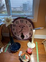 Image of large LG clock, tall amber vase with artificial flower, decorative bowl, red candle and white desk lamp on hardwood floor by window