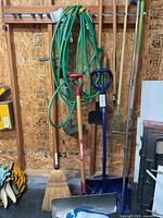 Overall view of garden tools hanging on plywood wall including garden hoses, shovels, broom, snow shovel, rake