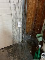 Photo showing nine wire plant cages in storage against a wall, made of metal wire with circular rings and vertical supports.