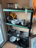 Front angled view of the 4 tier black plastic shelf filled with various kitchenware items such as glass bowls, metal mixing bowl, oven mitts, plates and trays.