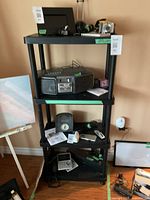 Front view of black plastic shelving unit with various electronic items and accessories placed on shelves for display purposes, which are excluded from sale.