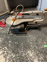 Ryobi 16" variable speed scroll saw resting on a textured concrete floor with power cord visible and several blade packets nearby.