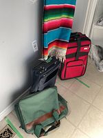 Four travel items standing against the wall: red wheeled suitcase, smaller blue carry-on bag, green travel bag, and colorful throw blanket visible hanging.
