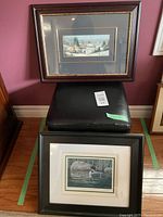 Photo showing two framed prints and a black faux leather storage stool stacked in front of a wall. The upper print depicts a winter village scene and is framed in dark wood. The lower print depicts a loon with chick in a black frame.