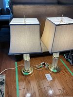 Two contemporary style table lamps with square fabric shades and cylindrical bases on a wooden floor in front of a couch.