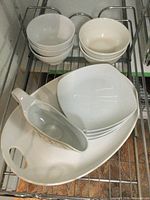 Photo showing white oval serving platter, white gravy boat, square plates, and small bowls stacked on metal rack.